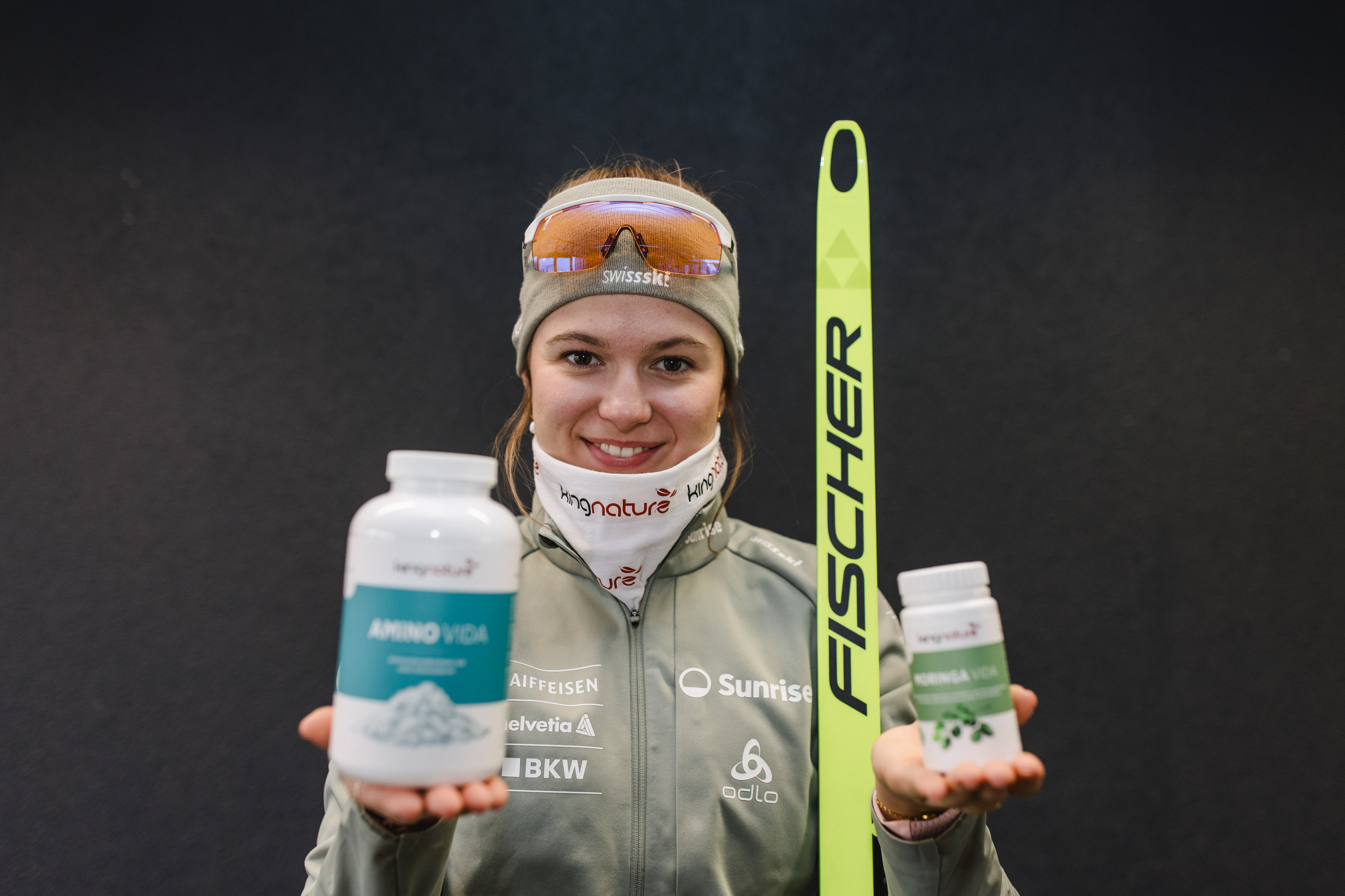 Eine Sportlerin, Ramona Schöpfer, in Wintersportkleidung lächelt, hält einen großen und einen kleinen Ergänzungsbehälter vor sich, neben ihr steht ein grüner Fischer-Ski vor einem dunklen Hintergrund.