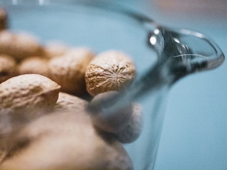 Nahaufnahme ganzer Muskatnusssamen in einem Glasbehälter vor einem sanften blauen Hintergrund. Der Fokus liegt auf einer aromatischen Muskatnuss, während mehrere andere im Hintergrund unscharf sind.