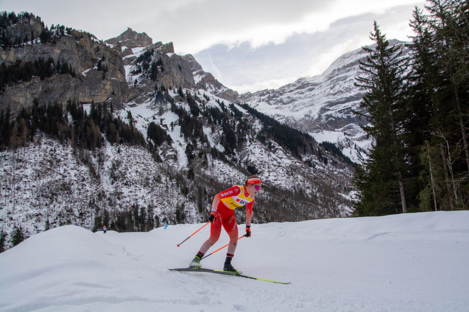 Ramona Schöpfer, in auffallend roter Kleidung, rast über eine schneebedeckte Piste, umrahmt von hohen Kiefern und schroffen, schneebedeckten Bergen unter einem bewölkten Himmel.