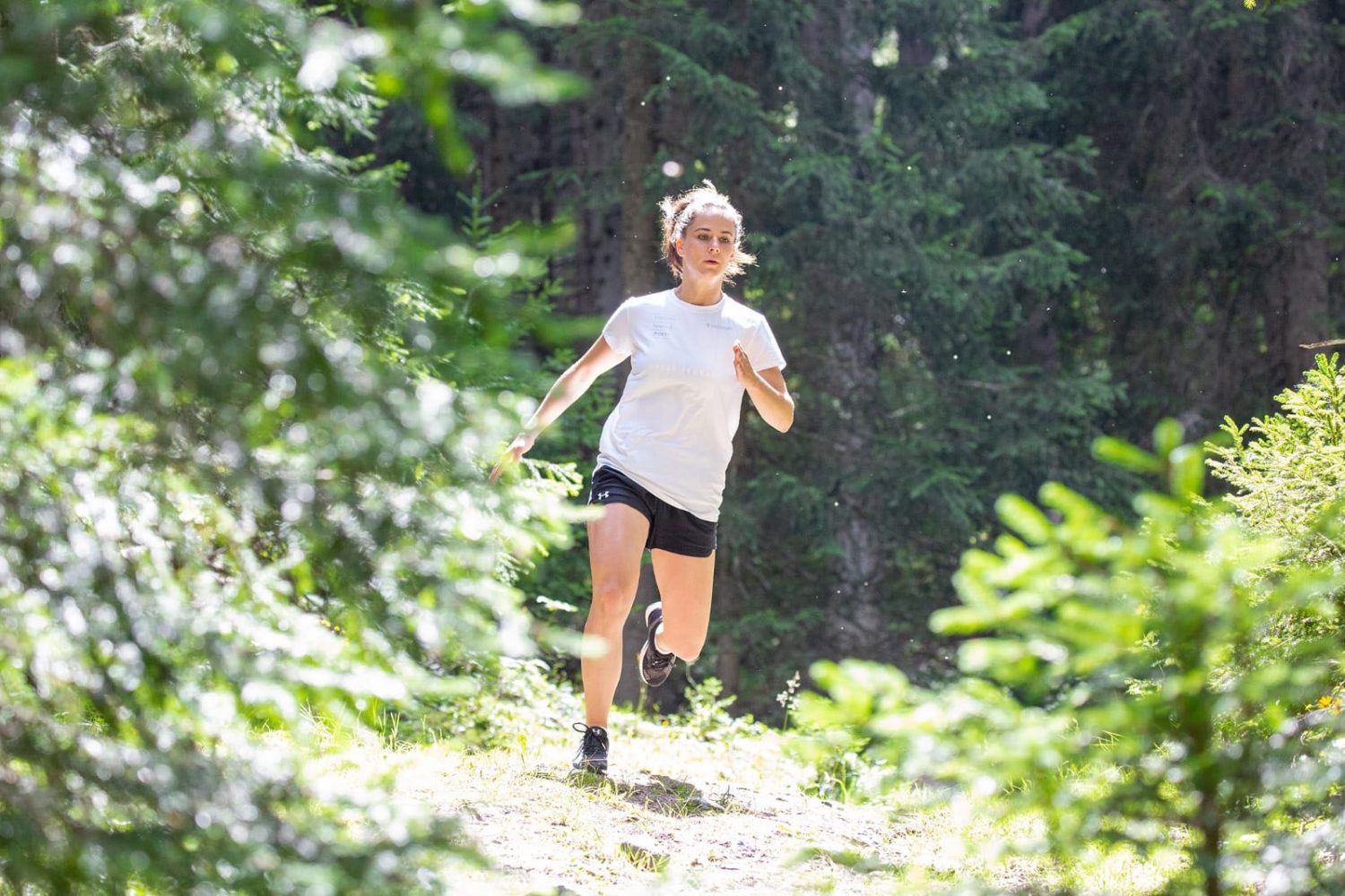 Talina Gantenbein läuft in einem weißen Shirt und schwarzen Shorts durch einen sonnenbeschienenen Waldweg, umgeben von üppigen grünen Bäumen und Laub.