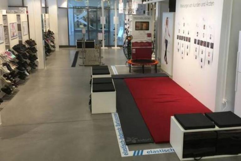 Ein modernes Ladeninterieur mit grauem Boden, weißen Wänden und Regalen mit Kybun-Schuhen. Ein roter Teppich verläuft in der Mitte, mit Bänken und einem kleinen Trampolin am Ende nahe einer Ausstellungswand im Center Arbon-Hamel.