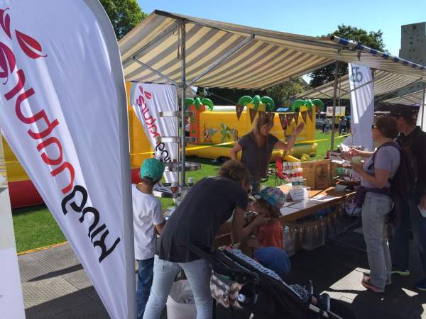Menschen treffen sich am Stand des Wädilaufs 2017 unter einem gestreiften Vordach. Eine Frau, Vertreterin von kingnature, steht hinter einem Tisch mit Broschüren und Artikeln, während Kinder und Erwachsene miteinander interagieren. Im Hintergrund sind eine aufblasbare Attraktion und Bäume zu sehen.