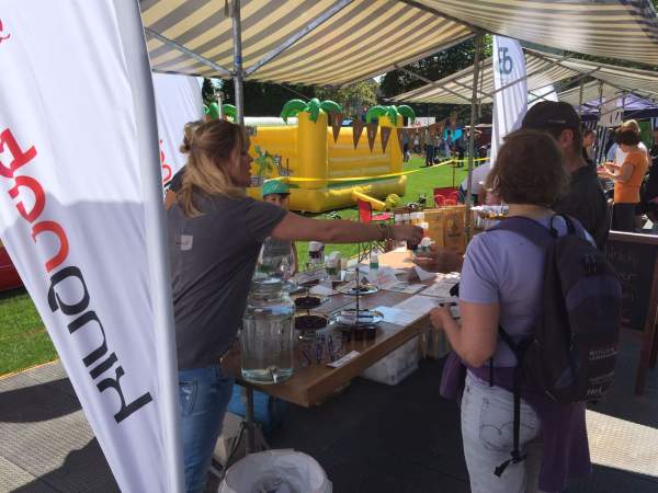 Eine Frau reicht einem Besucher an einem Außenstand unter einem gestreiften Baldachin beim Wädilauf 2017 ein Getränk. Die Leute versammeln sich um den Tisch, im sonnigen Hintergrund sind eine gelbe Hüpfburg und das Logo von kingnature zu sehen.