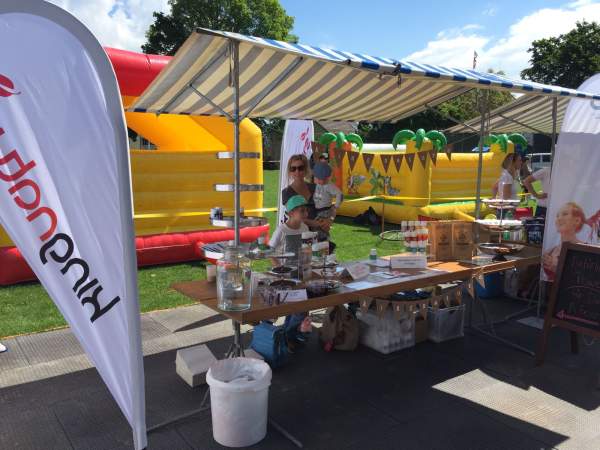 Eine Frau und zwei Kinder stehen hinter einem Tisch mit Speisen und Getränken auf dem Wädilauf 2017. Im Hintergrund sind an diesem sonnigen Tag Banner, ein gestreiftes Vordach und ein gelbes aufblasbares Spielgerät zu sehen.