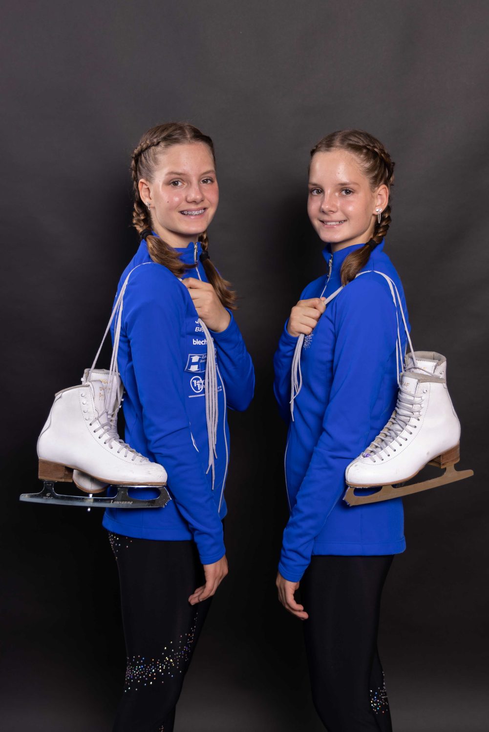 Selina Arnold und Alessia Arnold, zwei junge Mädchen mit geflochtenen Haaren, stehen lächelnd nebeneinander in passenden blauen Jacken und schwarzen Hosen. Jede trägt ein Paar weiße Schlittschuhe über der Schulter vor einem schlichten, dunklen Hintergrund.