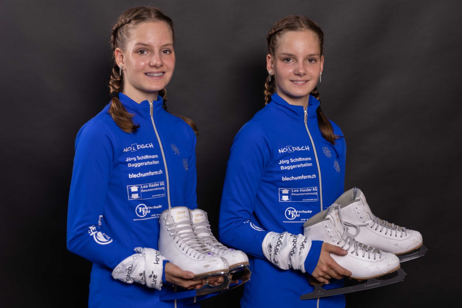 Selina Arnold und Alessia Arnold, zwei junge Eiskunstläuferinnen in passenden blauen Jacken, stehen Seite an Seite, halten weiße Schlittschuhe und posieren vor einem dunklen Hintergrund.