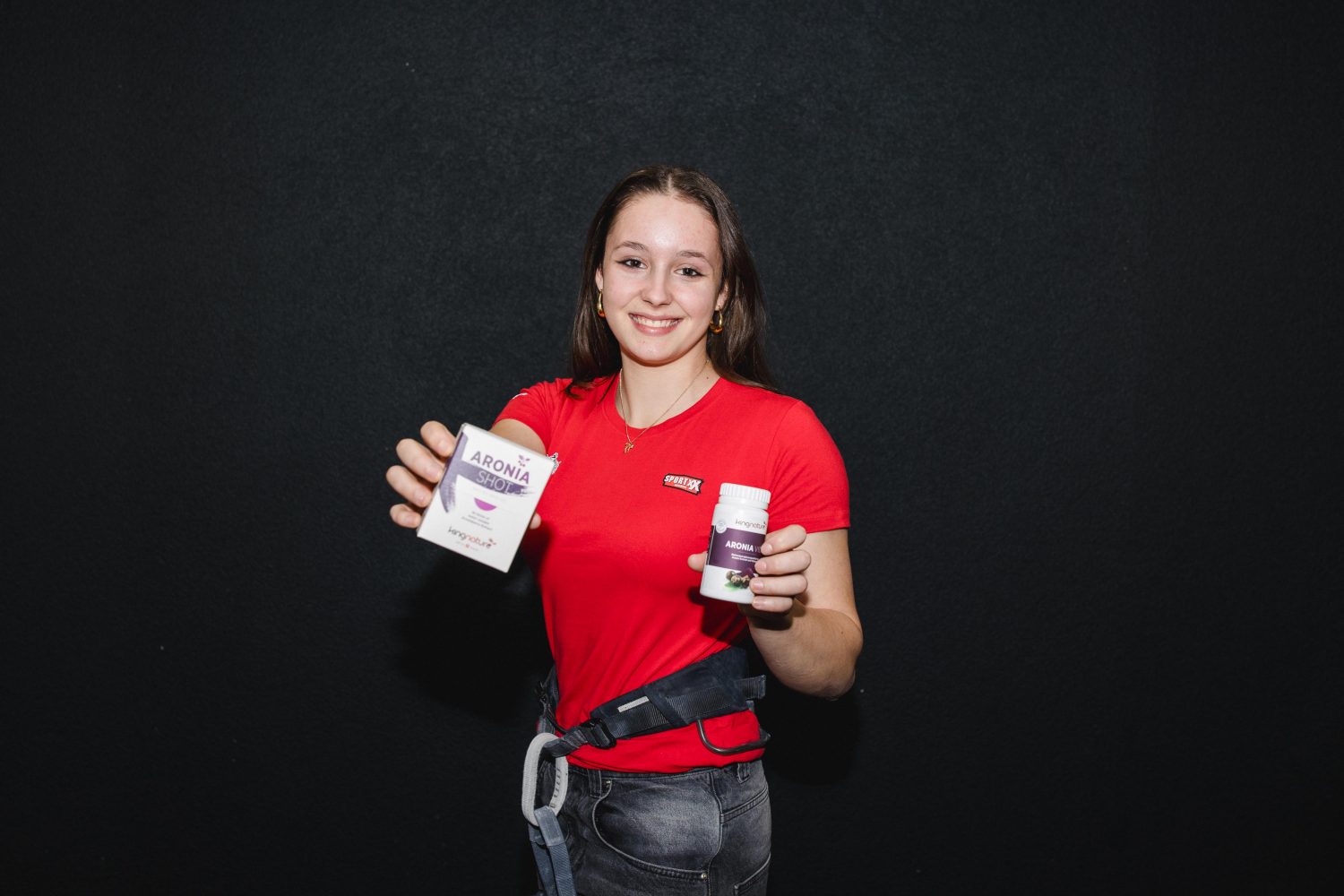 Valentina von Gunten, eine junge Frau im roten Hemd, lächelt und hält vor einem dunklen Hintergrund eine Schachtel und eine Flasche Aronia-Nahrungsergänzungsmittel hoch.