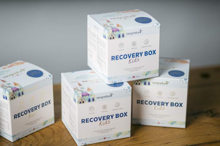 Vier weiße Boxen mit der Aufschrift „RECOVERY BOX Kids“ und farbenfrohen Häuserillustrationen sind auf einer Holzfläche gestapelt. Helle Symbole und ein verspielter Text deuten darauf hin, dass es sich um ein ansprechendes Unterstützungsprodukt für Kinder handelt.