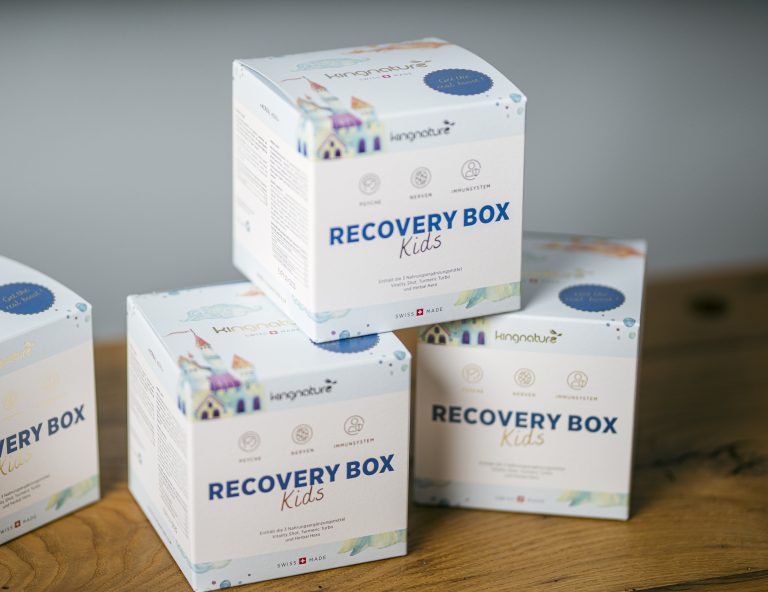 Vier weiße Boxen mit der Aufschrift „RECOVERY BOX Kids“ und farbenfrohen Häuserillustrationen sind auf einer Holzfläche gestapelt. Helle Symbole und ein verspielter Text deuten darauf hin, dass es sich um ein ansprechendes Unterstützungsprodukt für Kinder handelt.