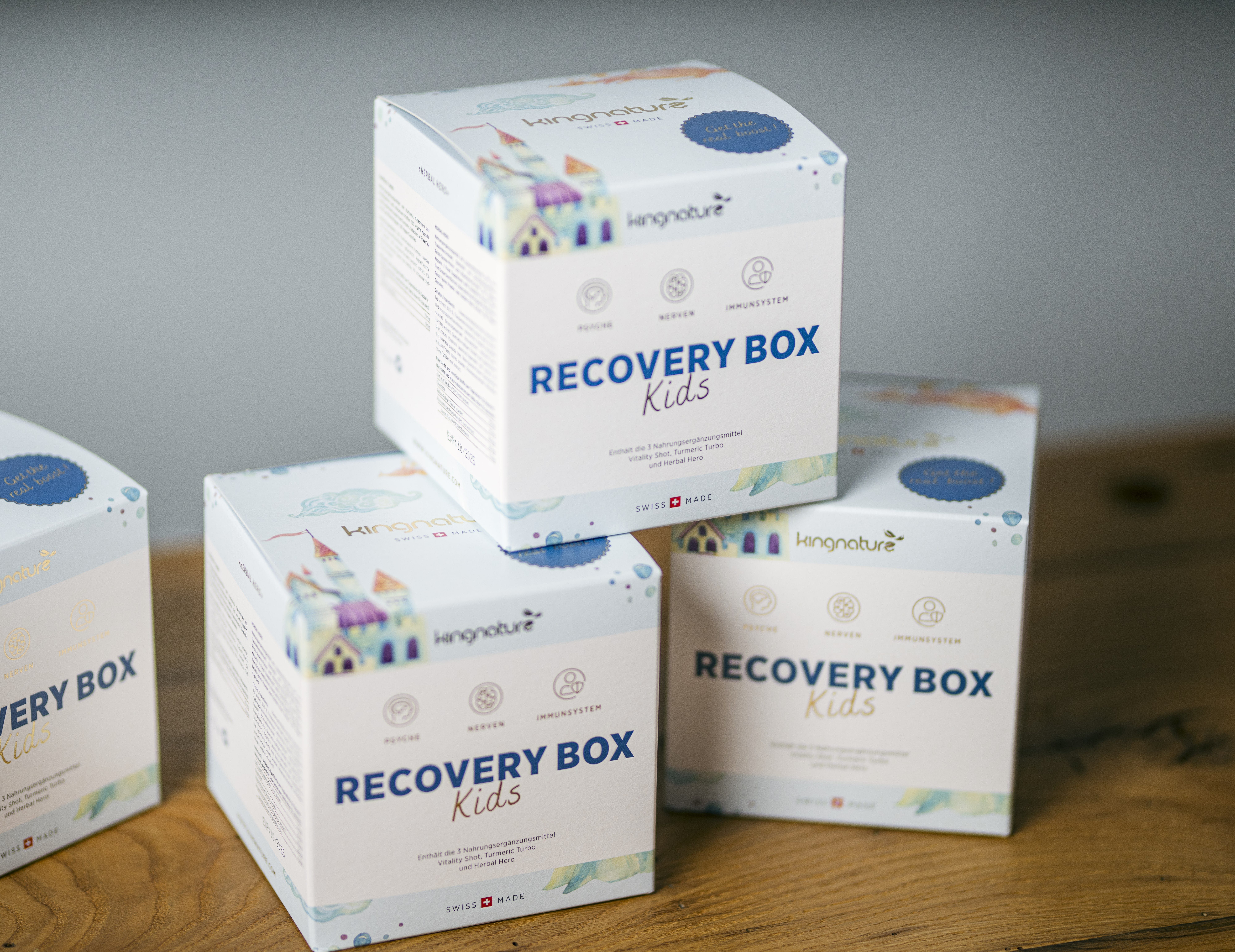 Vier weiße Boxen mit der Aufschrift „RECOVERY BOX Kids“ und farbenfrohen Häuserillustrationen sind auf einer Holzfläche gestapelt. Helle Symbole und ein verspielter Text deuten darauf hin, dass es sich um ein ansprechendes Unterstützungsprodukt für Kinder handelt.