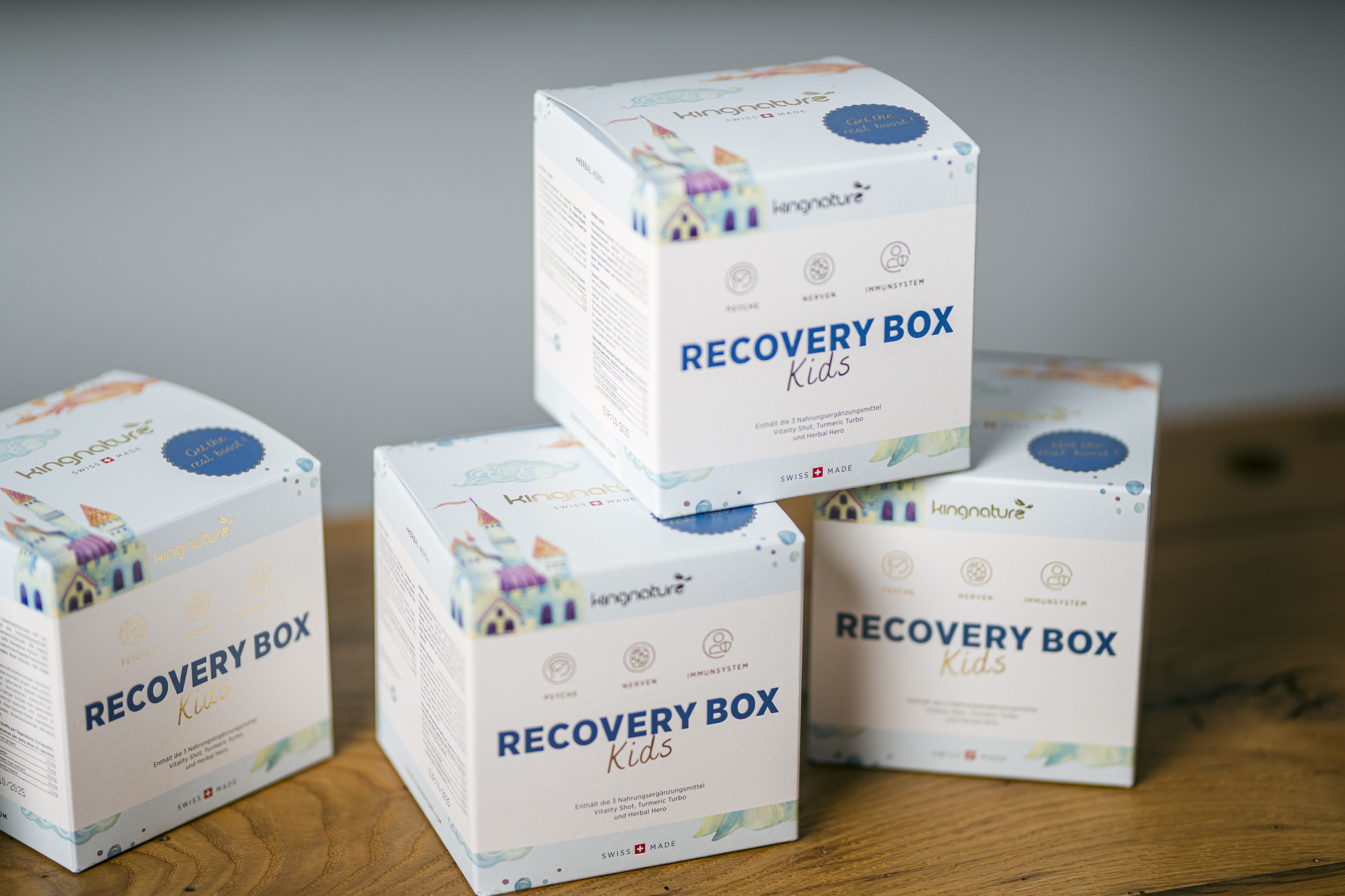 Vier weiße Boxen mit der Aufschrift „RECOVERY BOX Kids“ und farbenfrohen Häuserillustrationen sind auf einer Holzfläche gestapelt. Helle Symbole und ein verspielter Text deuten darauf hin, dass es sich um ein ansprechendes Unterstützungsprodukt für Kinder handelt.