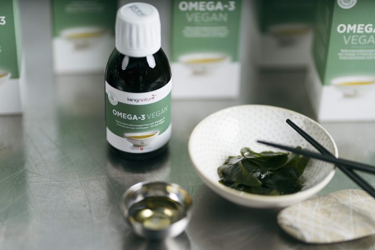 Eine Flasche mit einem veganen Omega-3-Nahrungsergänzungsmittel steht auf einer Metalloberfläche, neben einer kleinen Schale mit Seetang und Essstäbchen, einer kleinen Schale mit Öl und im Hintergrund Schachteln mit veganen Nahrungsergänzungsmitteln.