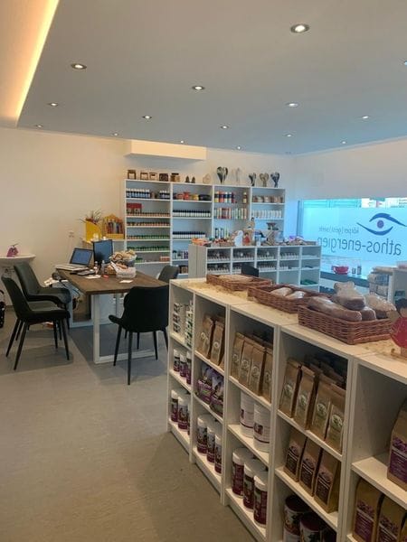 Ein heller, moderner Wellness-Shop mit Produkten von Athos-Energien. In den Regalen werden Nahrungsergänzungsmittel präsentiert, und ein Schreibtisch mit zwei Computern steht für Beratungen bereit. Tageslicht fällt durch ein Fenster mit Logo.
