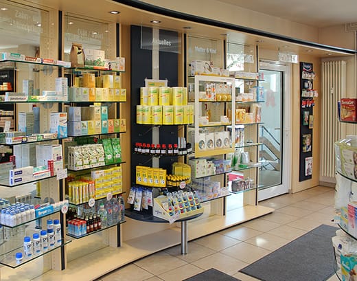 In den Regalen einer gut beleuchteten Westend Apotheke werden verschiedene Gesundheitsprodukte, darunter Schachteln, Flaschen und Verpackungen, präsentiert. Im Hintergrund sind eine Glastür und ein Fenster zu sehen. Der Boden ist gefliest.