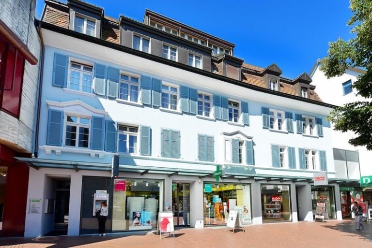 Ein dreistöckiges Gebäude mit blauen Fensterläden und Ziegeldach, mit mehreren Ladenfronten wie der Drogerie Brugg, breiten Fenstern und Glastüren auf Straßenebene an einem sonnigen Tag.