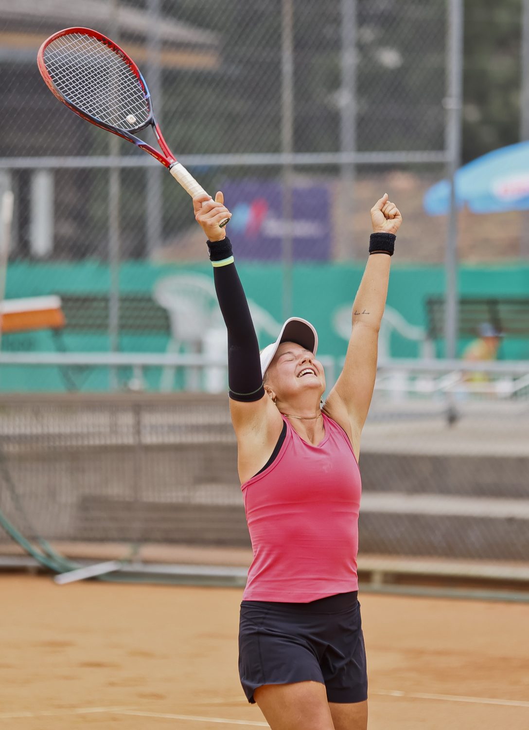 A female tennis player in a pink top and black shorts raises her arms and tennis racket in celebration on a clay court, smiling joyfully with eyes closed.