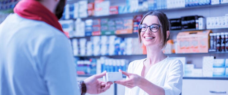 Ein lächelnder Apotheker mit Brille in der AVIE Apotheke in der Sachsen-Allee überreicht einem Kunden über den Apothekentresen hinweg eine kleine weiße Schachtel. Im Hintergrund sind Regale mit Produkten zu sehen.