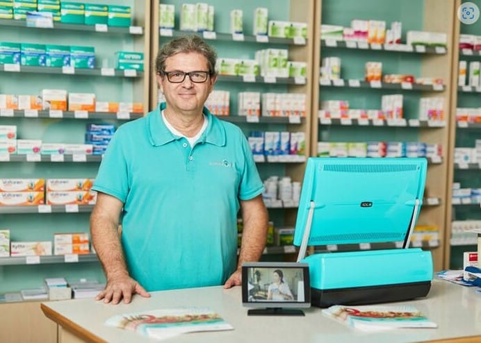 Ein Apotheker in einem türkisfarbenen Hemd steht hinter dem Tresen der St. Hubertus Apotheke, umgeben von Regalen mit verschiedenen Apothekenartikeln und Rezeptboxen. Auf dem Tresen liegen eine leuchtend blaue Kasse und Werbematerialien.