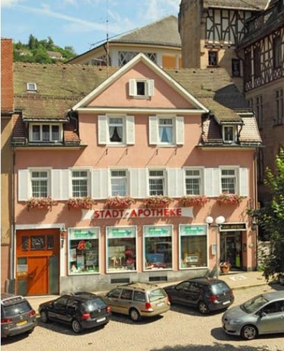 Ein rosafarbenes, dreistöckiges Gebäude mit weißen Fensterläden beherbergt die Stadt-Apotheke. Davor parken auf einer Kopfsteinpflasterstraße unter sonnigem Himmel mehrere Autos.