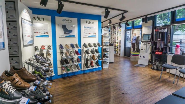 Die Inneneinrichtung eines Schuhgeschäfts der upswing GmbH zeichnet sich durch Holzböden, Regale mit Sport- und Freizeitschuhen, Informationsplakate an den Wänden und natürliches Licht aus, das durch große Fenster hereinströmt. Ein Sockenständer und ein Sitzbereich sind ebenfalls zu sehen.