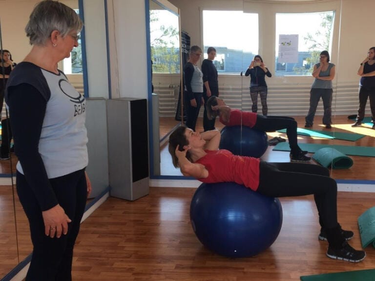 Regula Gehrig im roten Shirt macht Sit-ups auf einem blauen Gymnastikball, während ein Trainer in einem hellen Fitnessstudio mit Spiegeln und anderen Personen im Hintergrund zusieht.