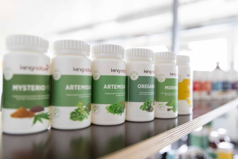 Flaschen mit Nahrungsergänzungsmitteln mit grün-weißen Etiketten, darunter Produkte mit den Bezeichnungen Hysteria, Artemisia, Oregano und Moringa, stehen in einem Regal in einem gut beleuchteten Raum aufgereiht. Preisanpassungen ab März 2023 sind eventuell relevant.