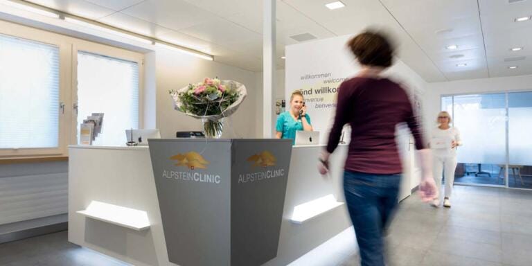Der Empfangsbereich einer modernen Gesundheitsklinik zeigt eine Mitarbeiterin hinter dem Tresen, einen Blumenstrauß auf dem Tresen und zwei Frauen, die durch die Lobby gehen. Auf dem Tresen prangt das Logo der Alpstein Clinic AG.
