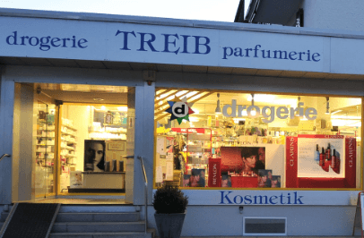 Im Eingangsbereich eines hell erleuchteten Ladens mit großen Schaufenstern sind Regale voller Schönheits- und Hygieneprodukte zu sehen. Über dem Eingang stehen Schilder mit den Aufschriften „Drogerie“, „Treib“, „Parfümerie“ und „Kosmetik“.