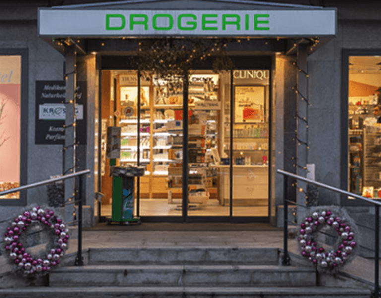 Ein Ladeneingang zur Kressig Drogerie mit Glastüren, auf beiden Seiten mit Ornamenten geschmückte Weihnachtskränze und im Inneren unter warmer Beleuchtung sichtbare Regale mit Produkten.