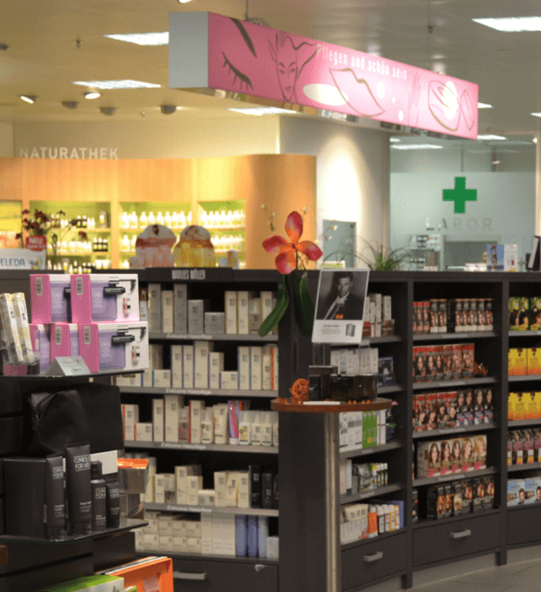 Regale mit verschiedenen Hautpflege- und Schönheitsprodukten in einer gut beleuchteten Drogerie. Darüber hängt ein rosa Schild mit Abbildungen von Gesichtern und Make-up-Artikeln, während eine dekorative Blume einen kleinen Tisch im Gang schmückt.