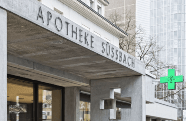 Ein Betongebäude mit dem Schriftzug „Apotheke Süßbach“ und einem grün beleuchteten Apothekenkreuz mit Schlangensymbol auf der rechten Seite; modernes, urbanes Ambiente.