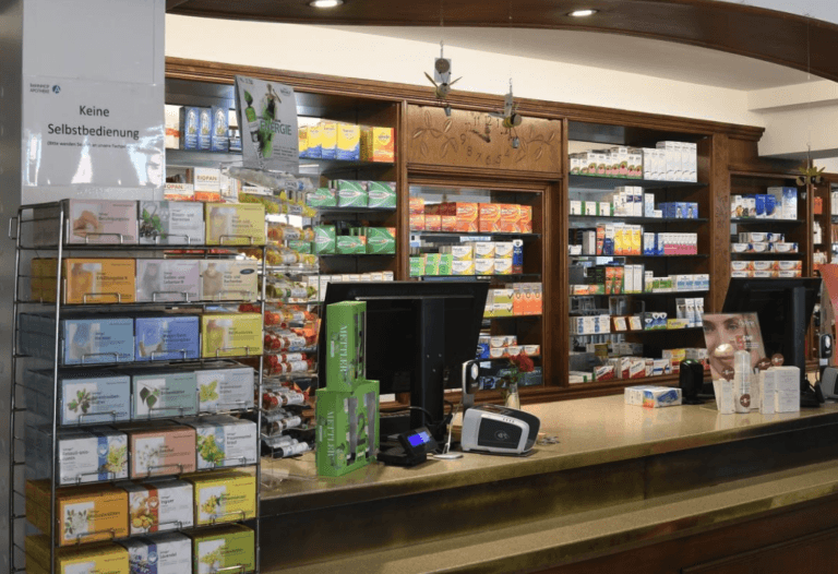 Ein Apothekenschalter in der Bahnhof Apotheke in Wetzikon ist mit Regalen mit Medikamentenboxen und Gesundheitsprodukten, zwei Computerbildschirmen und einem Kartenzahlungsterminal ausgestattet. Links steht auf einem Schild „Keine Selbstbedienung“.