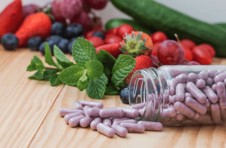 Ein Glasgefäß mit violetten Kapseln ergießt sich auf eine Holzoberfläche, umgeben von frischen Früchten wie Erdbeeren, Weintrauben, Blaubeeren und Gurken sowie Minzblättern – eine Inspiration für Wohlbefinden durchs Leben und Wohlfühlprodukte.