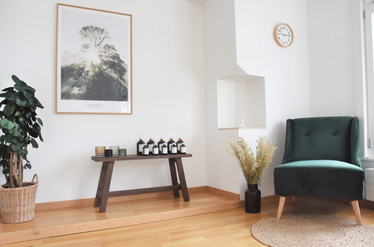 Ein gemütliches, minimalistisches Zimmer von Manuela Moshayedi mit einem grünen Sessel, einer kleinen Bank mit Flaschen und Kerzen, einer Topfpflanze, einer Wanduhr, einem großen Kunstwerk mit Naturmotiven und getrockneten Blumen in einer schwarzen Vase auf dem Holzboden für ein natürliches Ambiente.