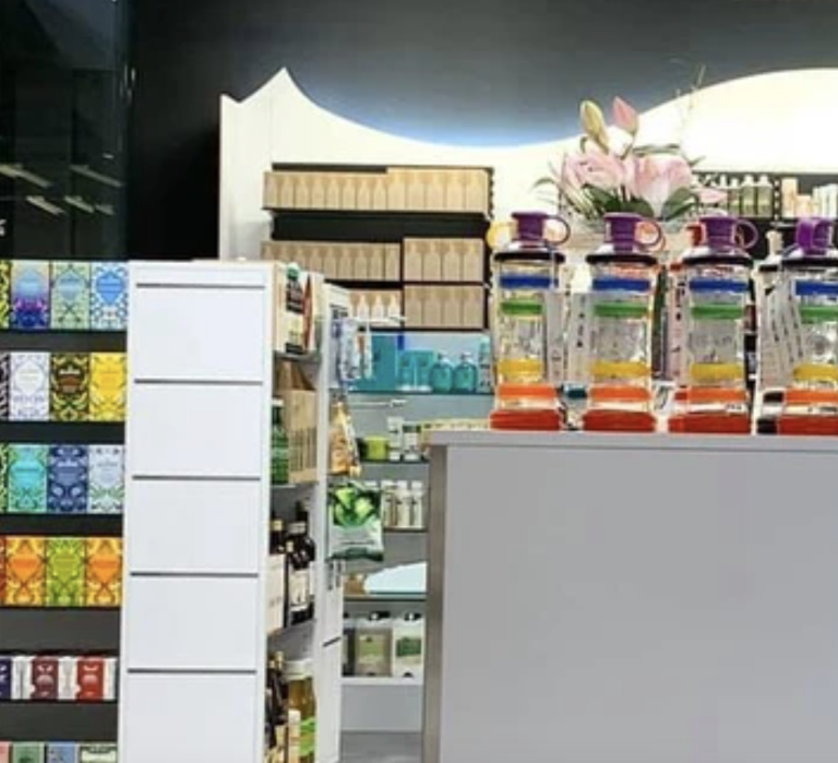 Im Ladeninneren sorgen bunte Flaschen auf einer Theke, Regale voller Kartons und verschiedener Produkte sowie ein farbenfrohes Blumenarrangement im Hintergrund für eine einladende Atmosphäre.