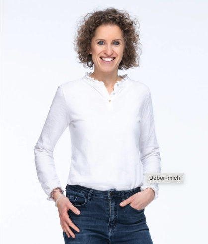 Eine lächelnde Frau mit lockigem Haar, die eidgenössisch diplomierte Naturheilpraktikerin Raffaela Lehmann, trägt eine weiße Bluse und blaue Jeans und steht mit einer Hand in der Tasche vor einem schlichten weißen Hintergrund.