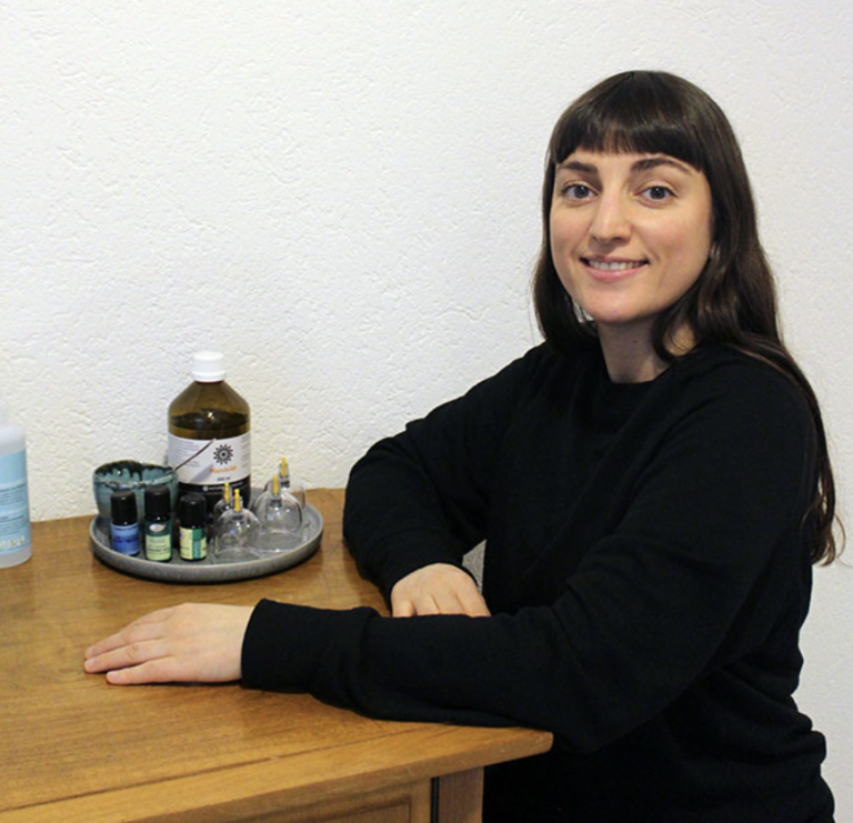 Janine Städler, mit glattem braunem Haar und Pony, sitzt an einem Holztisch in ihrer Naturheilpraxis. Vor ihr liegt ein Tablett, gefüllt mit Flaschen, Gläsern und Fläschchen mit Ölen und Tinkturen.