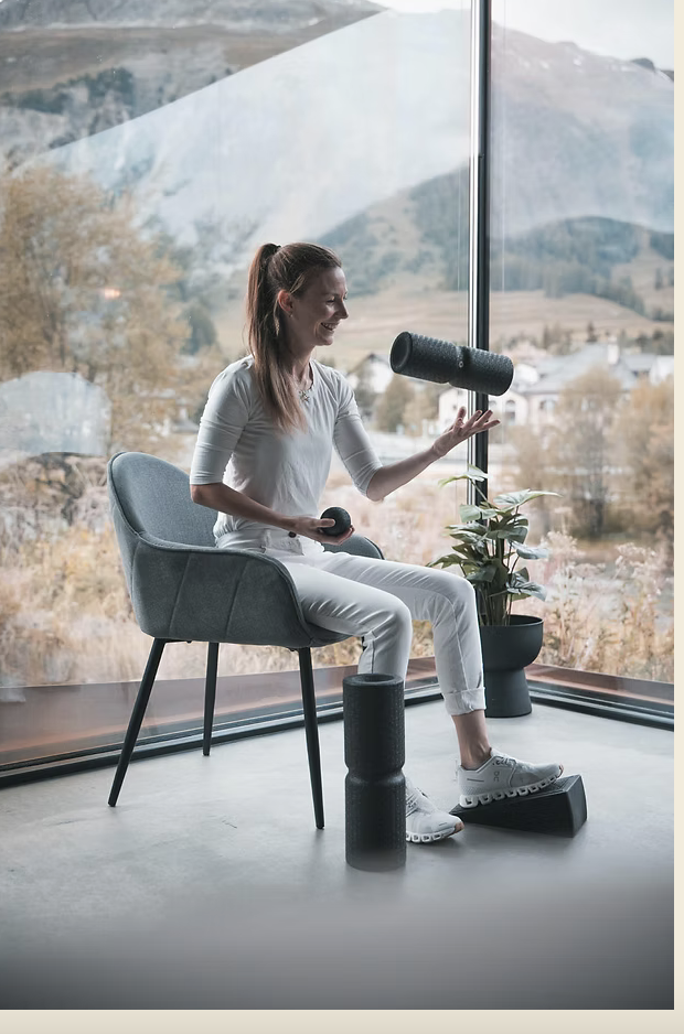 Deborah Rauch sitzt drinnen in Weiß und lächelt, während sie Schaumstoffrollen auf ihren Beinen balanciert und eine in der Hand hält. Große Fenster hinter ihr geben den Blick auf eine malerische Berglandschaft frei, die die ruhige Atmosphäre von Terapia Rauch unterstreicht.