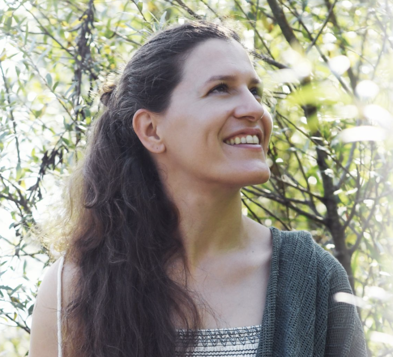 Eine Person mit langen braunen Haaren blickt lächelnd nach oben, umgeben von grünem Laub und Sonnenlicht. In der Naturheilpraxis Albis trägt sie ein gemustertes Oberteil und ein grünes Tuch, das über eine Schulter drapiert ist und den ganzheitlichen Wellness-Ansatz von Luzia Seeholzer verkörpert.