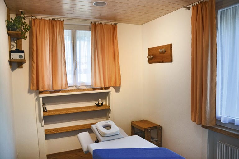 Ein gemütlicher Massageraum im MMP Trubschachen mit einem Massagetisch, der mit einem blauen Laken bedeckt ist, Holzregalen, orangefarbenen Vorhängen an den Fenstern und sanftem Tageslicht, das den Raum erfüllt.