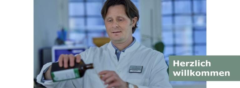 Ein Apotheker im weißen Kittel gießt in einem Labor der Drogerie Martinsplatz Flüssigkeit aus einer braunen Flasche. In der unteren rechten Ecke steht auf grünem Grund der Text „Herzlich willkommen“.