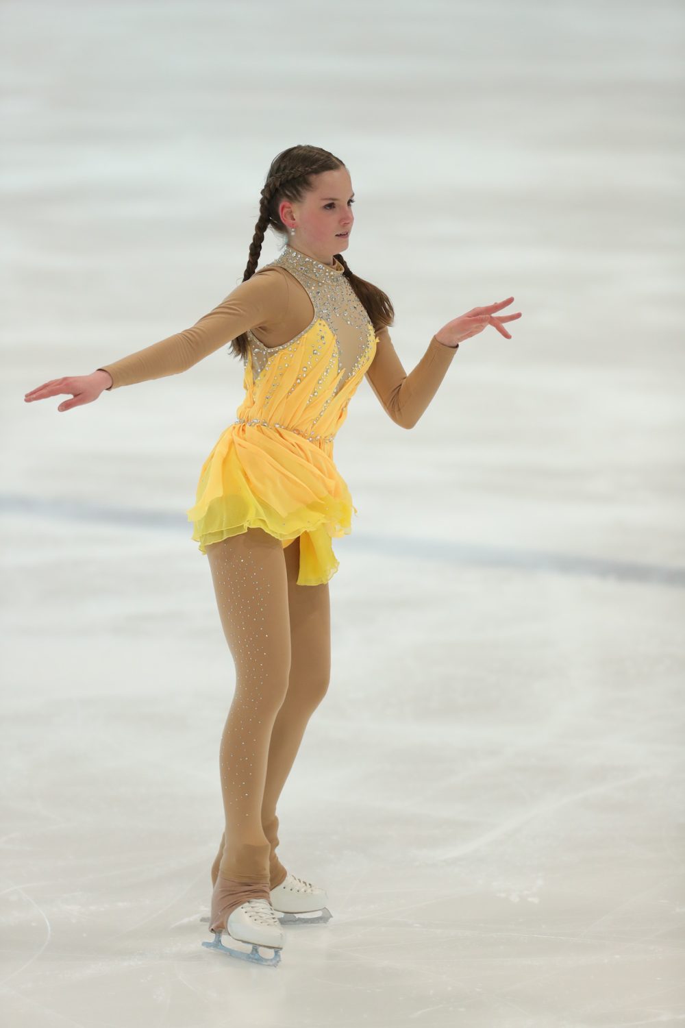 Alessia Arnold, eine Eiskunstläuferin in einem gelb-goldenen Kostüm mit Juwelenakzenten, gleitet mit anmutig ausgestreckten Armen und geflochtenem braunen Haar über das Eis.