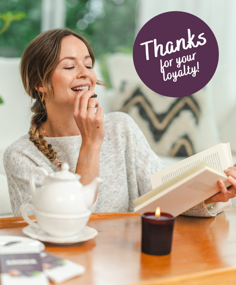 Eine lächelnde Frau sitzt mit einem Buch, einer Teekanne und einer brennenden Kerze an einem Tisch. Eine kreisförmige Grafik trägt in verspielter weißer Schrift den Text „Danke für Ihre Treue!“. Die gemütliche Szene zeigt kingnature, bekannt für Nahrungsergänzung aus der Schweiz.