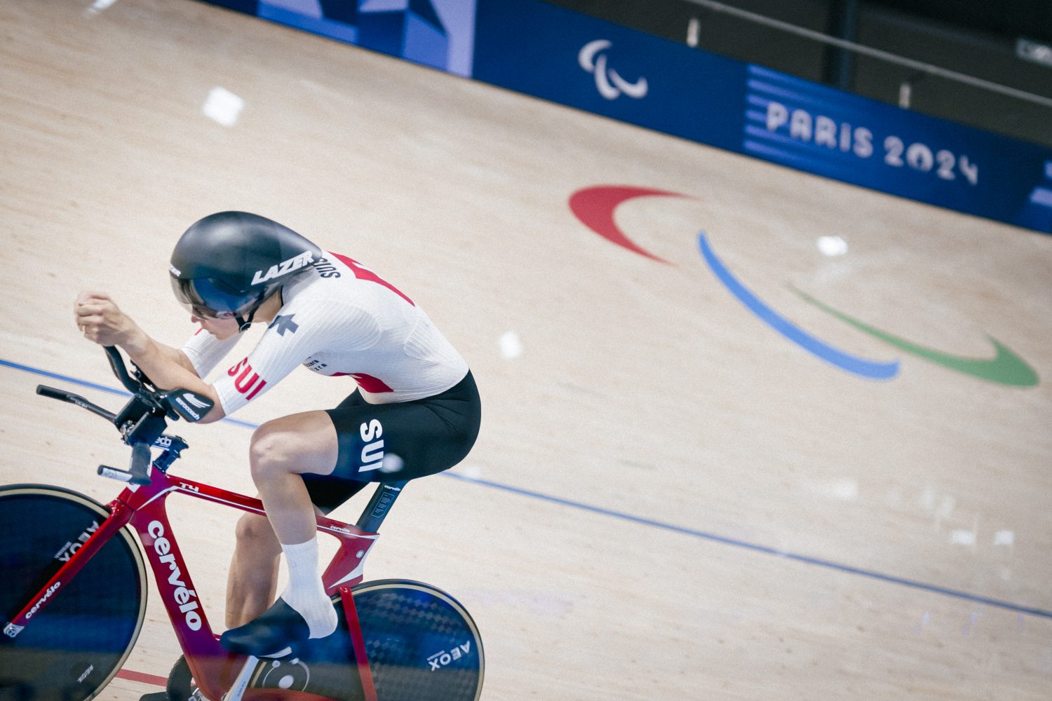 A cyclist wearing a white and red Swiss uniform races on a track bike at the Paris 2024 Paralympic Games, with the Paralympic symbol and PARIS 2024 visible in the background.