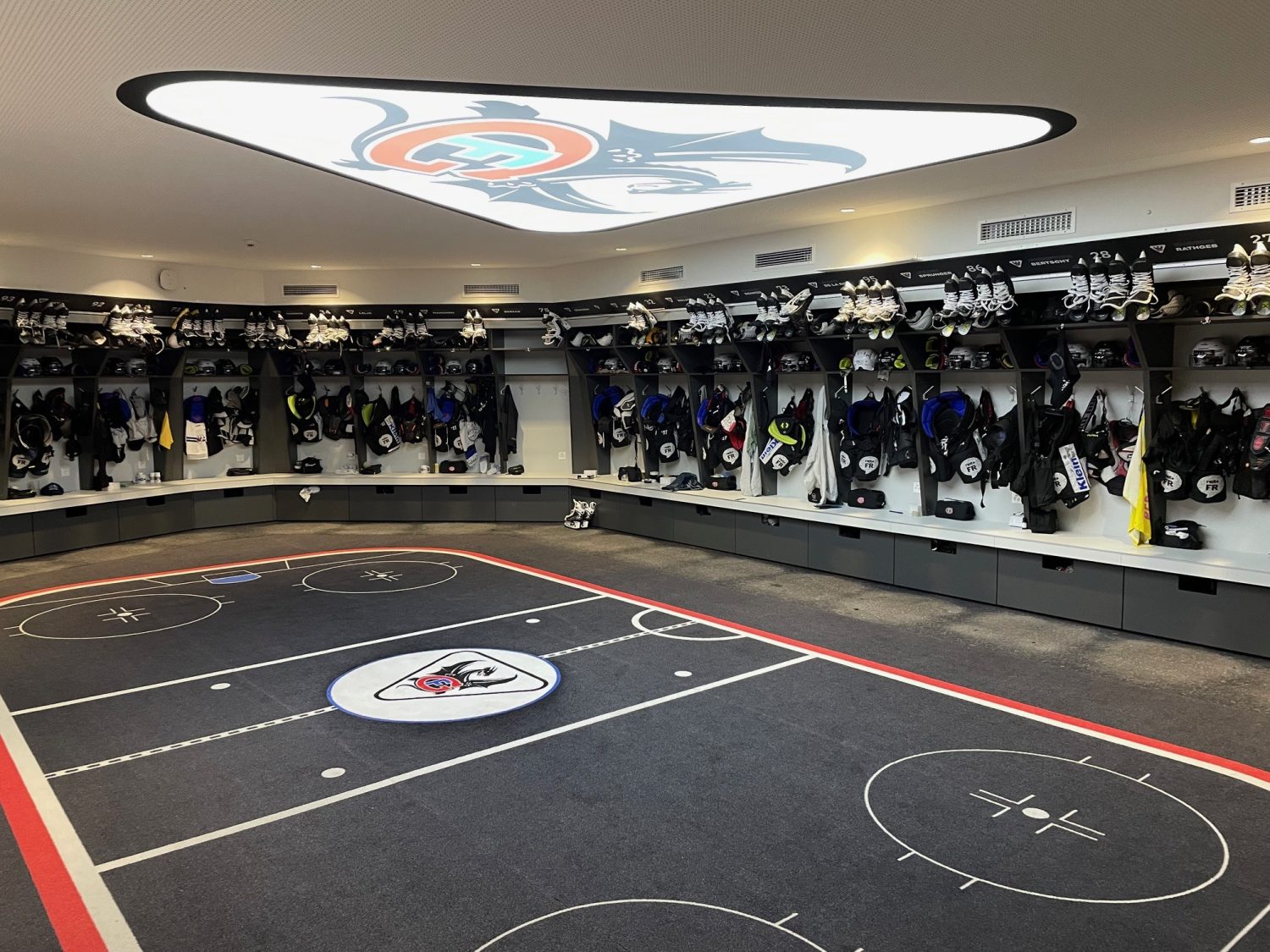 Eine geräumige, moderne Hockey-Umkleidekabine für den HC Fribourg-Gottéron. Ausrüstung und Uniformen sind ordentlich in Fächern entlang der geschwungenen Wände verstaut. Der Boden erinnert an ein Eishockeyfeld, und ein großes Teamlogo ziert die Decke – perfekt für Schweizer Eishockey.