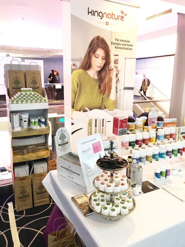 Ein Stand von kingnature in München präsentiert Nahrungsergänzungsmittel und Kosmetika, inspiriert von der Naturheilkunde. Bunte Flaschen, Broschüren und kleine Tiegel sind ordentlich auf Tischen und Regalen angeordnet, dahinter ein Banner mit einer lesenden Frau.