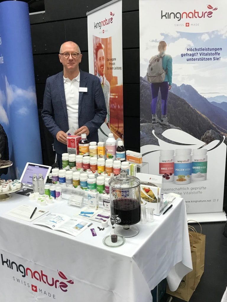 Ein Mann steht hinter einem Stand, der verschiedene Nahrungsergänzungsmittel und Wellnessprodukte von Kingnature auf dem Norddeutschen Heilpraktiker Kongress präsentiert. Auf dem Tisch liegen ein Poster mit einem Wanderer in den Bergen, Informationsbroschüren und ein Getränkeautomat.