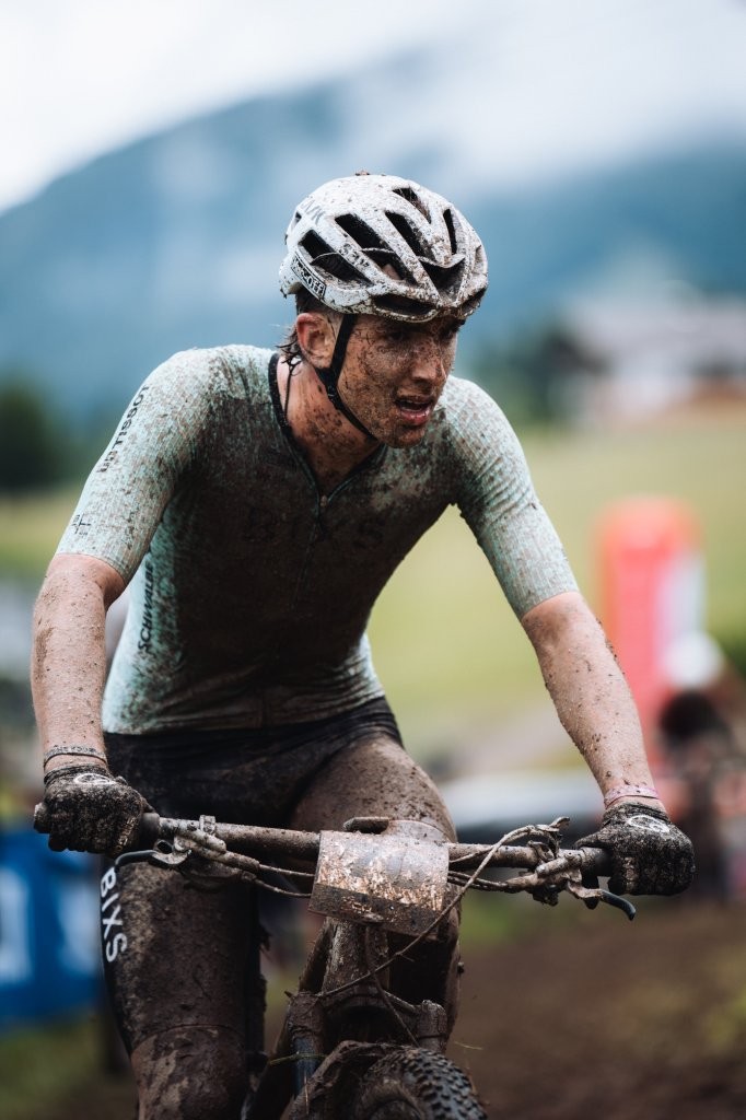 Ein mit Schlamm bedeckter Radfahrer fährt mit einem Mountainbike im Freien, trägt einen Helm und ein kurzärmeliges Trikot, mit einer verschwommenen Naturlandschaft im Hintergrund.