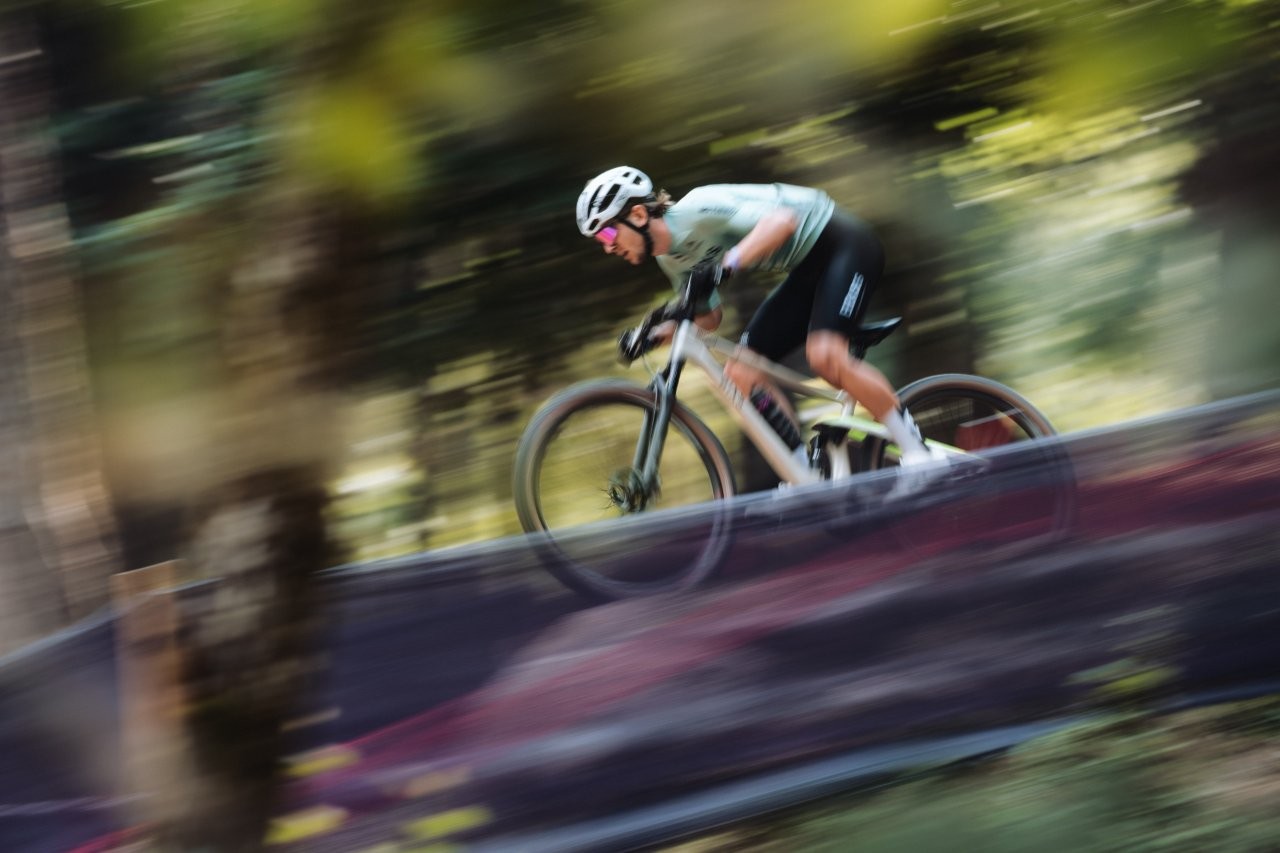 Ein Radfahrer, der einen Helm und eine Sonnenbrille trägt, fährt mit hoher Geschwindigkeit auf einem Mountainbike durch ein unscharfes, bewaldetes Gebiet, was Bewegung und Intensität suggeriert.