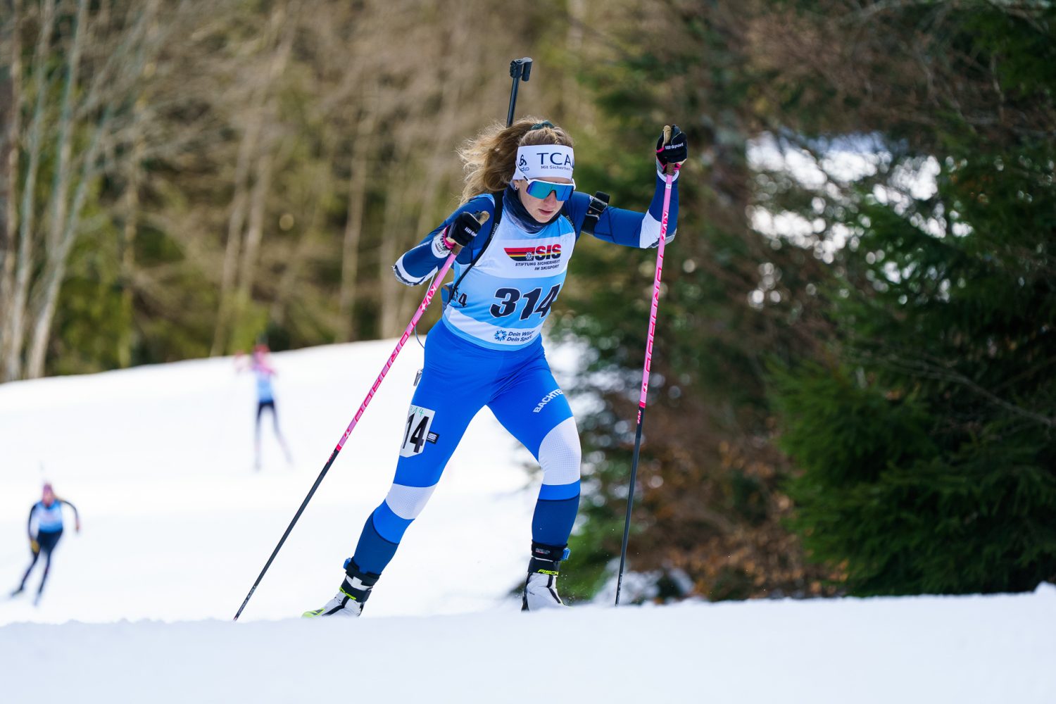 Eine Biathletin in blauer Kleidung und mit Sonnenbrille, identifiziert als Lisa Naegeli, fährt auf einer verschneiten Piste mit einem Gewehr auf dem Rücken bergauf; andere Skifahrer sind im Hintergrund in der Nähe von Bäumen zu sehen.